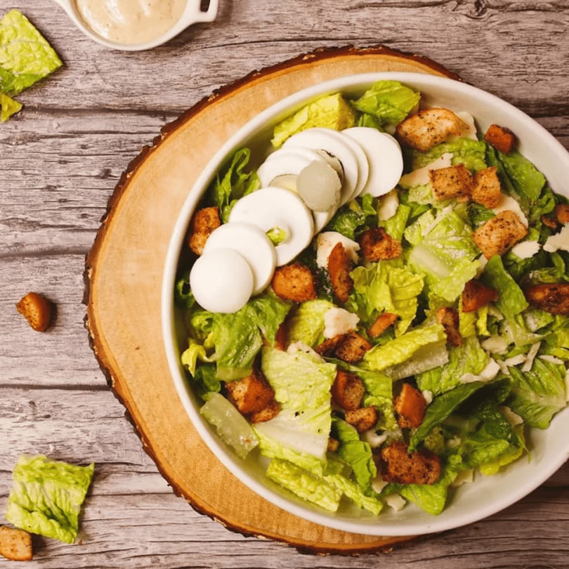 Caesar salad with lettuce, croutons, sliced hard-boiled eggs, and dressing on a wooden board.
