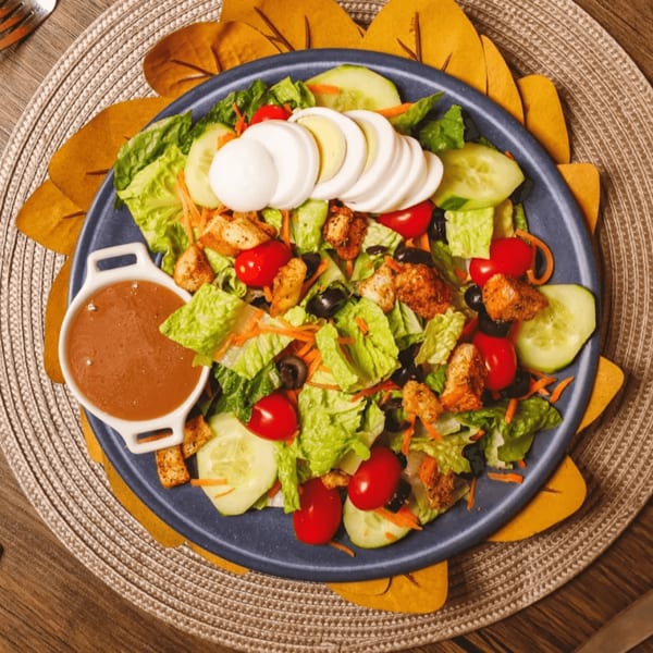 Chicken salad with lettuce, tomatoes, cucumbers, olives, hard-boiled eggs, and dressing on a blue plate.